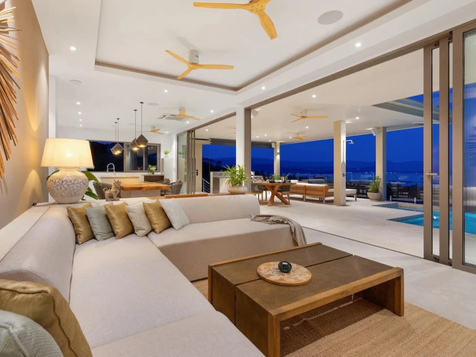 Salon intérieur le soir, grand canapé crème ouvert sur baies vitrées et piscine éclairée